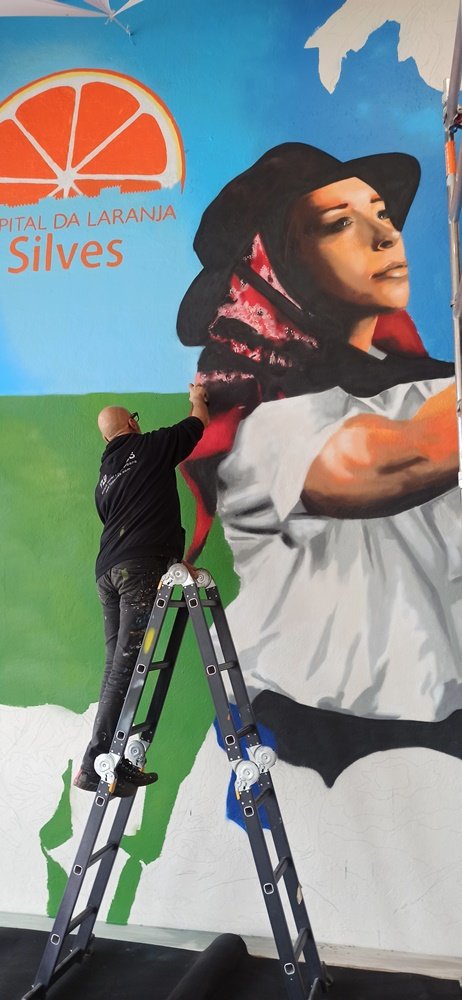 O artista Fly Pontes em cima do escadote a pintar pormenores no lenço da jovem. A parte superior da figura está quase pronta, mas ainda faltam pintar laranjas e folhagem na zona inferior. O logotipo do evento é visível.