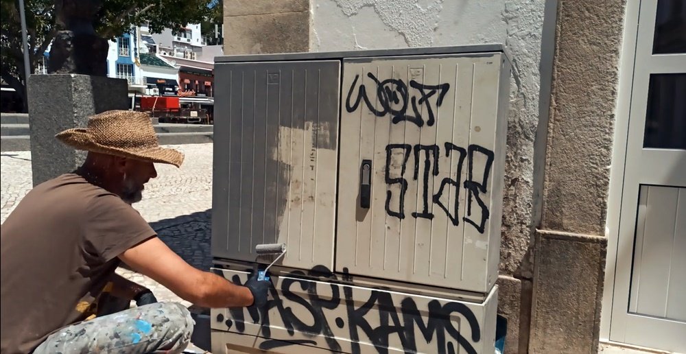 O artista Fly Pontes no local, a aplicar a tinta primária (primer) no armário de electricidade. As tags de spray no armário ainda são visíveis, marcando a transição da arte de rua para a pintura de homenagem no Largo da Meia Laranja.