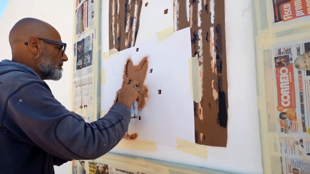 O artista Fly Pontes a pintar a primeira camada de stencil do gato. O artista usa um pauzinho para pressionar a folha, evitando que a tinta passe por baixo da folha. As cortinas da janela falsa já foram parcialmente pintadas.