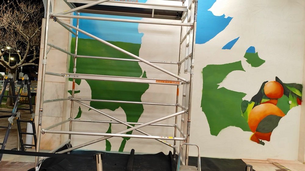 Resultado da pintura do primeiro dia do mural "Mostra da Laranja 2025". O azul do céu, o verde dos campos, e duas ou três laranjas com folhagem formam a silhueta da jovem. A base da composição em Silves.