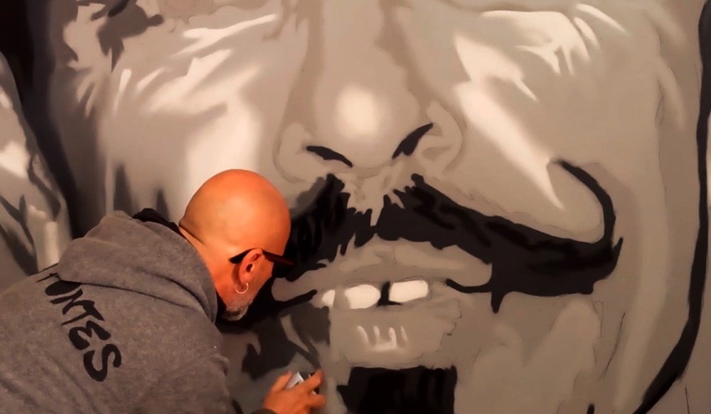 O artista Fly Pontes a aplicar os tons cinza na figura do retrato. A imagem mostra a progressão na modelagem, com os tons claros e escuros já visíveis, conferindo volume e realismo à figura que contrasta com o fundo azul.