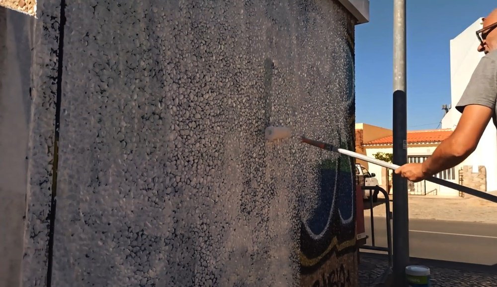 O artista Fly Pontes a aplicar o primário com um rolo na parede do PT de Almancil. Alguns graffiti antigos são visíveis por baixo da tinta, mostrando o processo de preparação da superfície para o novo mural.