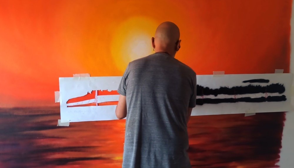 O artista Fly Pontes a pintar o stencil da silhueta da cidade de Portimão. O pôr do sol no fundo já está finalizado em tons vibrantes de laranja, criando o enquadramento ideal para o horizonte.