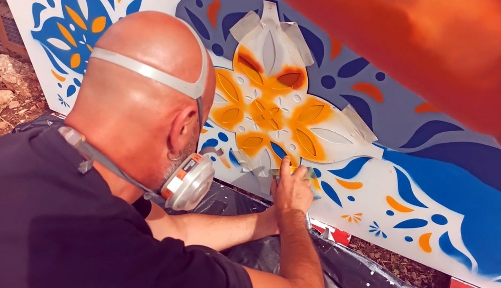 O artista Fly Pontes a pintar o stencil dos azulejos no mural "GeoPalcos". Vista aproximada da aplicação da máscara de stencil para replicar o padrão de azulejo, garantindo a geometria e a precisão da tradição portuguesa.