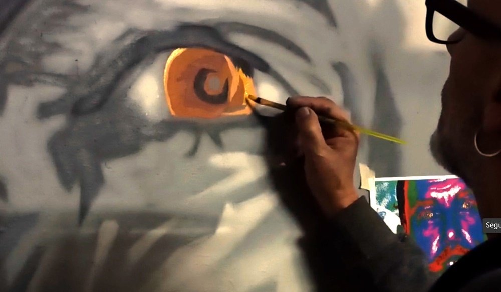 O artista Fly Pontes a pintar os olhos de castanho no retrato. A utilização do pincel fino permite a máxima precisão neste detalhe. A introdução da cor nos olhos é crucial para conferir vida e foco à figura principal em tons de cinza.