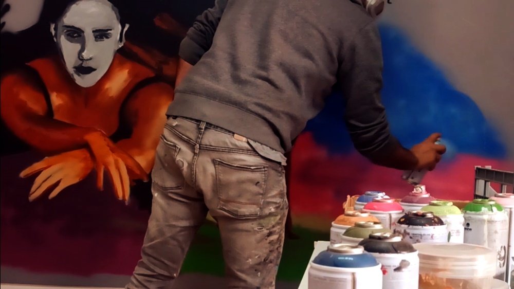 O artista Fly Pontes a criar os esfumados para a ilusão de fumo que envolve as bailarinas no mural. Em primeiro plano, latas de spray. Em segundo plano, uma das figuras da pintura a emergir deste efeito etéreo e místico de fumo.