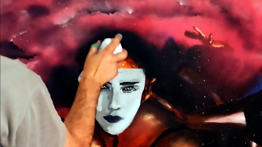 O artista Fly Pontes a aplicar esfumados por cima da bailarina principal, que usa uma máscara de gesso no rosto. Em segundo plano, uma mão de outra bailarina emerge do fumo, reforçando a ilusão e o mistério do mural "A Graça das Bailarinas".