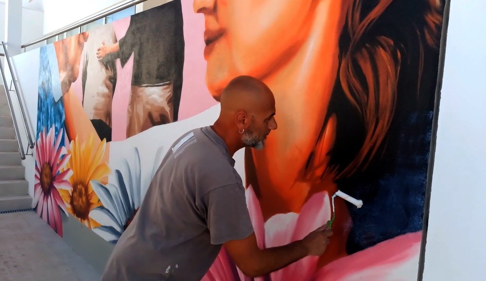 O artista Fly Pontes em primeiro plano a aplicar o verniz em todo o mural "Afecto e Cuidado em Lar de Idosos", já terminado. O verniz protege a obra e confere um acabamento brilhante e duradouro, selando a mensagem de afecto e acolhimento.