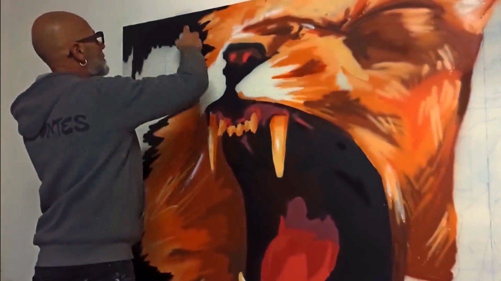 O artista Fly Pontes a pintar o fundo de preto no mural "Treino e Paixão". O leão estilizado já está quase concluído, emergindo do fundo escuro com as suas cores originais, criando um forte contraste.