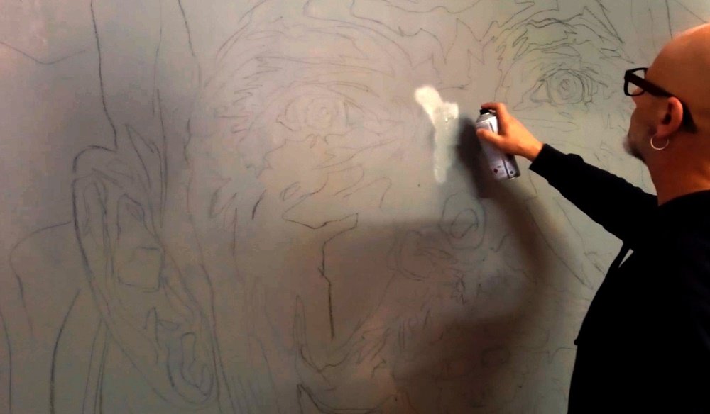 O artista Fly Pontes a aplicar os primeiros tons de cinza no retrato do mural ("De Estudo Efémero..."). A pintura está a ser guiada pelas linhas a carvão previamente traçadas, definindo a estrutura da figura principal em realismo monocromático.