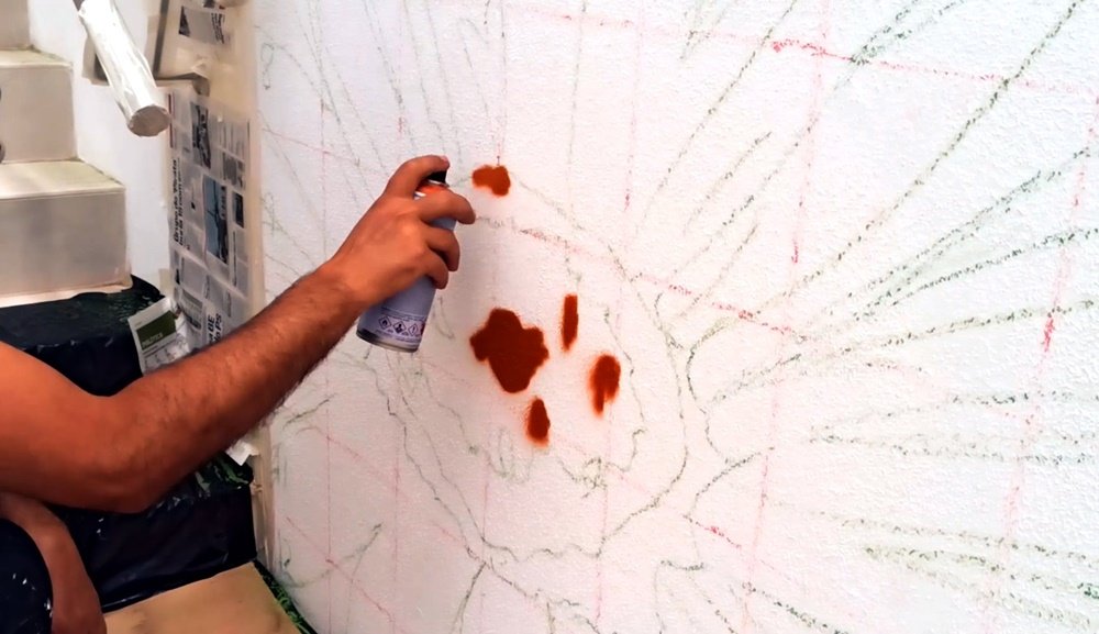 O artista Fly Pontes a iniciar a pintura de uma das flores em primeiro plano, no mural do Lar de Idosos. As linhas guia a carvão são visíveis, enquanto a lata de spray aplica os primeiros tons castanhos, definindo a forma do elemento floral.