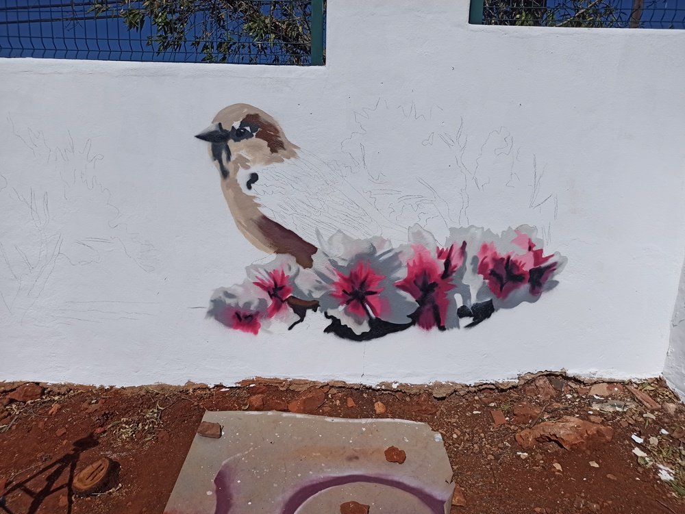 Início da pintura do mural "O Pardal no Despertar das Amendoeiras em Flor". Aplicação das primeiras cores castanhas do pardal e da base das flores da amendoeira (cores brancas/rosas), definindo os elementos centrais do mural.