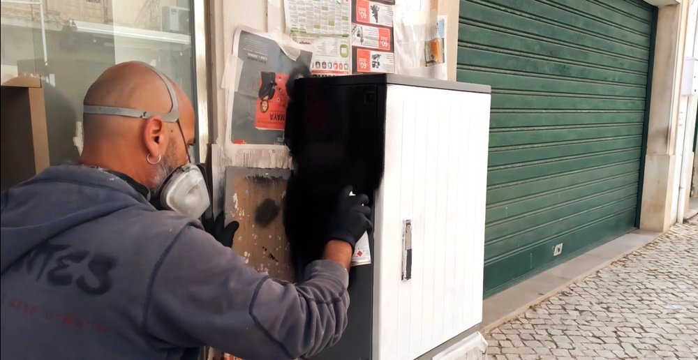 O artista Fly Pontes a pintar a lateral do armário com spray preto. A frente já está com primário branco, mostrando a preparação para um contraste estratégico no mural da Primeira Farmácia de Albufeira.