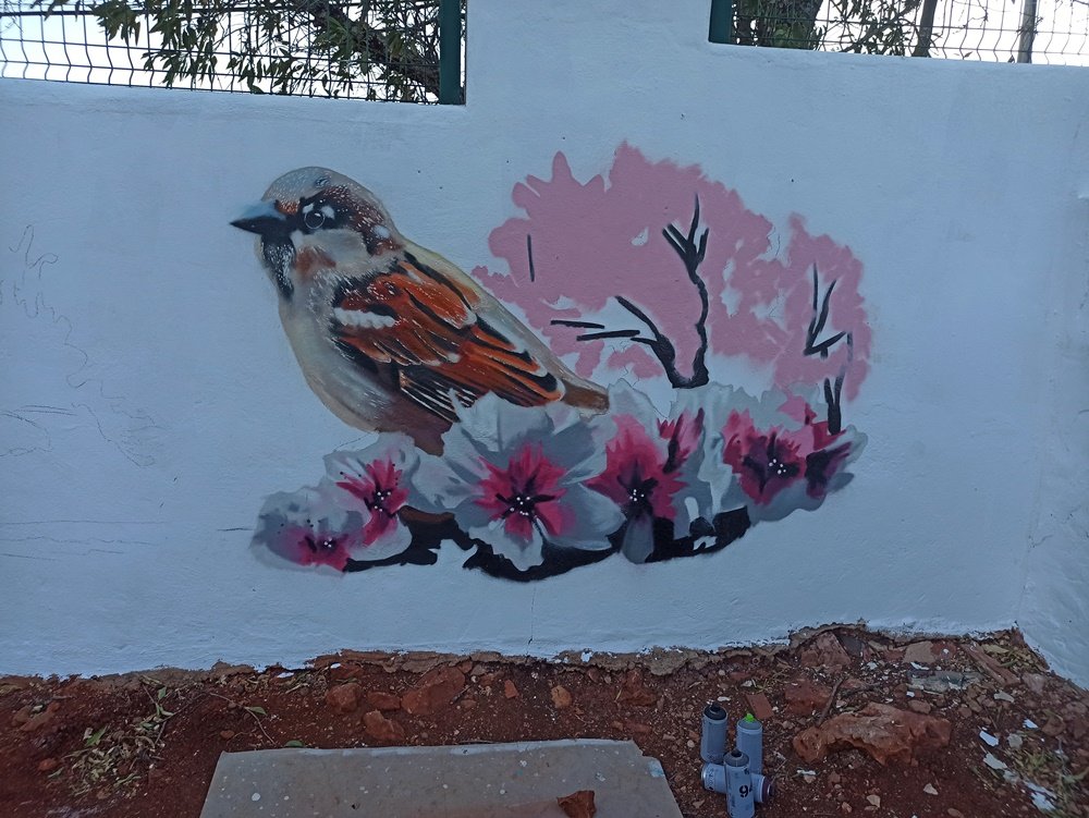 Fase avançada da pintura do mural Amendoeiras em Flor. O pardal e as flores de amendoeira em primeiro plano estão quase terminados. As primeiras cores da amendoeira mais afastada estão a ser aplicadas, criando o efeito de profundidade e perspectiva.