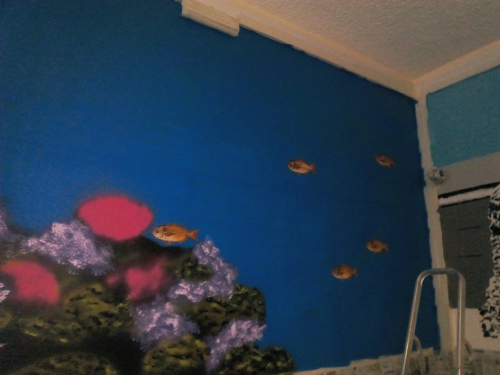 Processo do mural "O Ecossistema Marinho Local". Peixes pintados nadam em direcção ao coral em tons rosa e violeta. O fundo azul (água) ainda está em tons planos, sem modelagem de luz, e algumas rochas do coral estão incompletas.