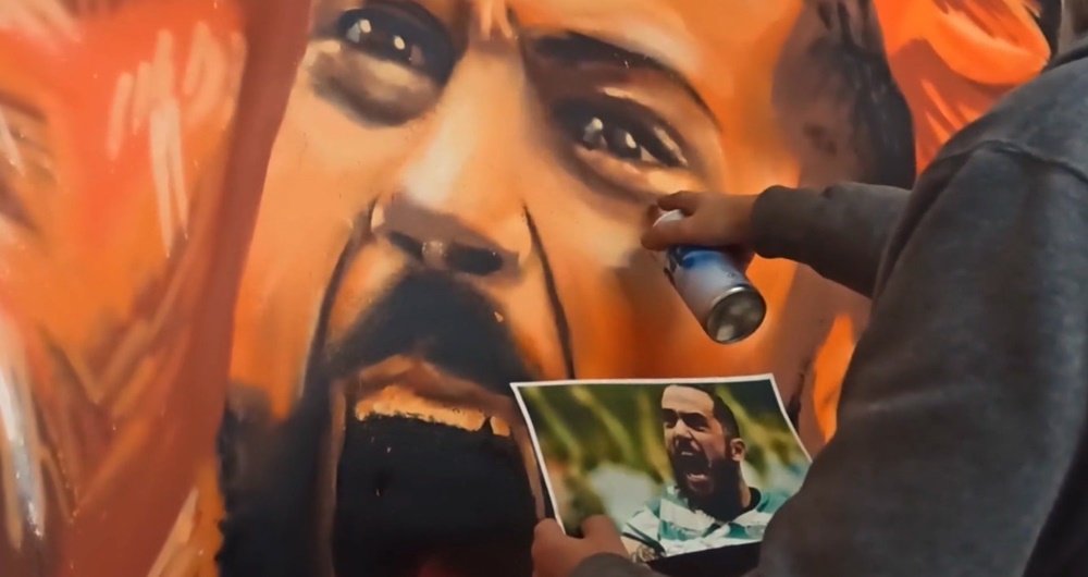 O artista Fly Pontes a pintar os olhos da figura de João Matos (Capitão Futsal Sporting CP) no mural. O artista segura a fotografia de referência na mão esquerda e a lata de spray na direita, focando-se no detalhe crucial que confere vida ao retrato.
