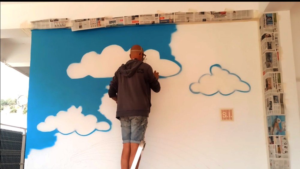 O artista Fly Pontes a pintar o céu de azul no mural "A Alegria de Pintar". O artista está a deixar espaços em branco estrategicamente, uma técnica que irá formar as nuvens, garantindo a luz e a leveza do fundo.