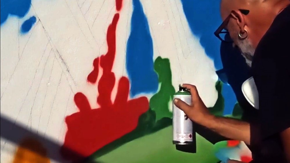 O artista Fly Pontes a pintar o verde da letra 'M' no mural "A Alegria de Pintar". A letra 'M' é parte do logótipo da Casa do Povo, de onde nasce simbolicamente o Arco-Íris, ligando visualmente a arte à identidade da instituição.