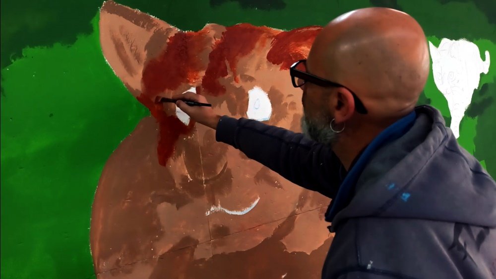 O artista Fly Pontes a pintar o Rato em grande plano no mural da Loja Petlandia. A aplicação dos tons castanhos está a modelar o volume e a textura do pelo do animal, parte essencial da narrativa de tensão com o Gato.