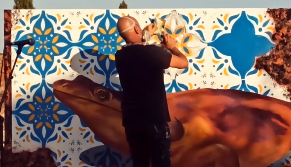 O artista Fly Pontes a pintar os azulejos no mural "GeoPalcos" (Geoparque Algarvensis). Vista afastada onde se vê o Metoposaurus Algarvensis quase pronto e a maioria dos painéis de azulejo concluídos. O mural funde geologia e tradição portuguesa.