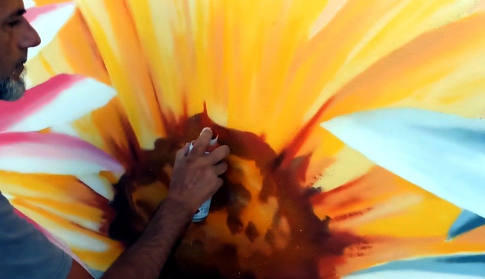 O artista Fly Pontes em grande plano a pintar os pormenores de uma das flores do mural do Lar de Idosos. A imagem destaca a precisão e o cuidado na aplicação dos acabamentos, que dão vida e realismo aos elementos florais.