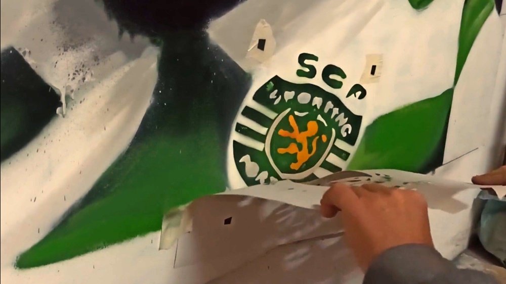 O artista Fly Pontes a remover a máscara de stencil do símbolo do Sporting CP no mural "Treino e Paixão". A remoção revela o emblema perfeitamente definido, contrastando com o retrato em spray livre.