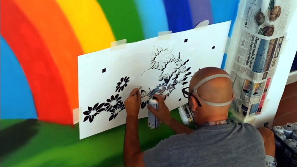 O artista Fly Pontes a pintar o stencil das flores no final do Arco-Íris, no mural "A Alegria de Pintar". A aplicação do stencil cria um campo de flores estilizadas, onde o Arco-Íris desagua, simbolizando a criatividade e a transição orgânica.