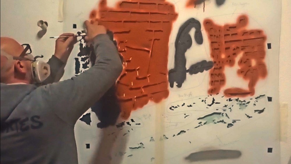 O artista Fly Pontes a pintar uma das camadas de stencil da grelha, no futuro mural da Julinha no Restaurante O Amândio. A Julinha ainda não está pintada, mostrando a construção da cena através do contexto da especialidade antes da figura principal.