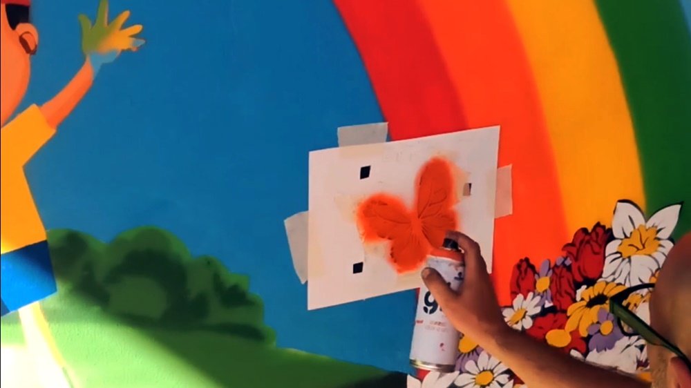 O artista Fly Pontes a pintar o stencil de uma das borboletas no mural "A Alegria de Pintar". As flores no final do arco-íris e parte de uma criança com as mãos sujas de tinta já estão visíveis, criando um contexto de criatividade e liberdade.