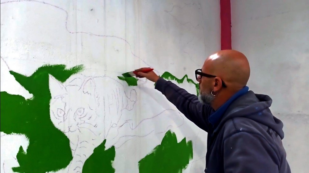 O artista Fly Pontes a pintar o fundo de verde no mural da Loja Petlandia. As linhas guias do Gato já são visíveis, indicando a futura localização e forma do animal, que irá emergir do vibrante fundo de tinta de água e acrílicos.