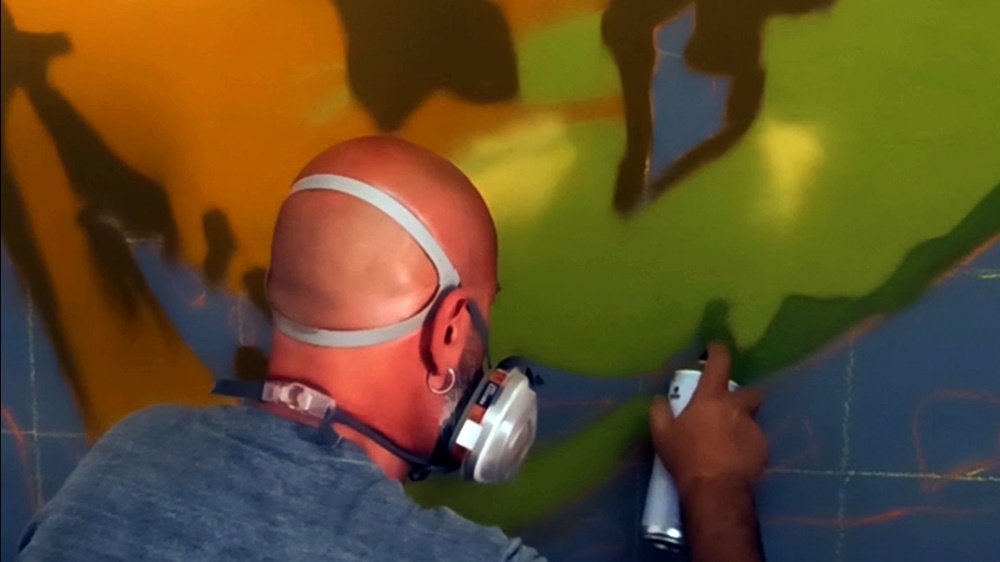 O artista Fly Pontes a aplicar os tons de verde no rosto do retrato. A quadrícula e as linhas guia são ainda visíveis, mostrando a progressão da experimentação de cor na figura principal antes do contraste final com o fundo violeta.