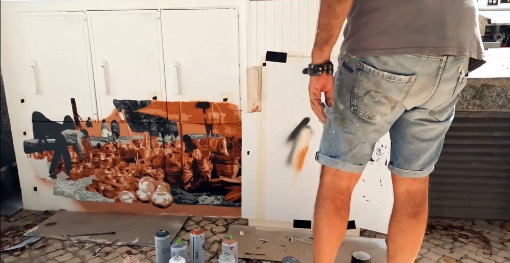 O artista Fly Pontes, de pé, a inspeccionar a pintura da Feira Franca de Albufeira. A imagem das pessoas em tons de cinza e os artigos da feira em tons castanhos já estão visíveis, confirmando o forte contraste visual e nostálgico.