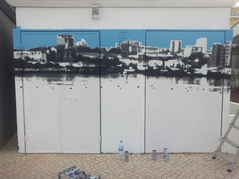 Todas as camadas de stencil do skyline de Portimão estão prontas, em contraste com o céu azul do mural "Homenagem aos Pescadores". Latas de spray no chão. A base paisagística está completa, aguardando os pescadores e o barco.