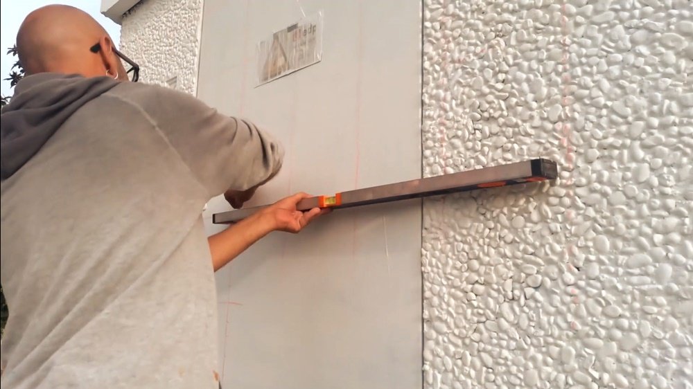 Traçado das linhas da quadrícula na frente do Posto de Transformação (PT) em Almancil. O artista utiliza um nível para garantir a precisão das linhas, que são marcadas com lápis de carvão, preparando o mural da Alfarroba e Agricultura.