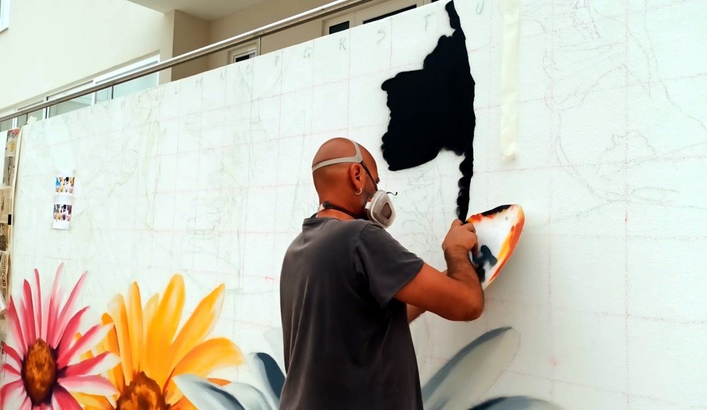O artista Fly Pontes em primeiro plano a pintar de preto a roupa de uma das figuras do mural. As flores em primeiro plano estão pintadas. Vê-se a quadrícula (método de grade) na parede não pintada, guiando a transferência do desenho em grande escala.