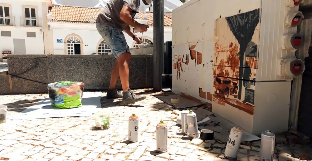O artista Fly Pontes no local, vai aplicar o spray sobre uma máscara de stencil colada no armário. Várias latas de spray em primeiro plano realçam a técnica de arte urbana em uso para a pintura da Feira Franca.