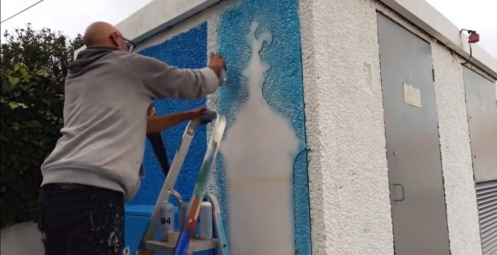 Pintura do fundo azul na lateral do PT em Almancil, para o mural da Chaminé Centenária e Andorinha. A andorinha já está pintada e a chaminé está coberta com uma máscara de stencil, preparando-se para receber a cor principal do fundo.