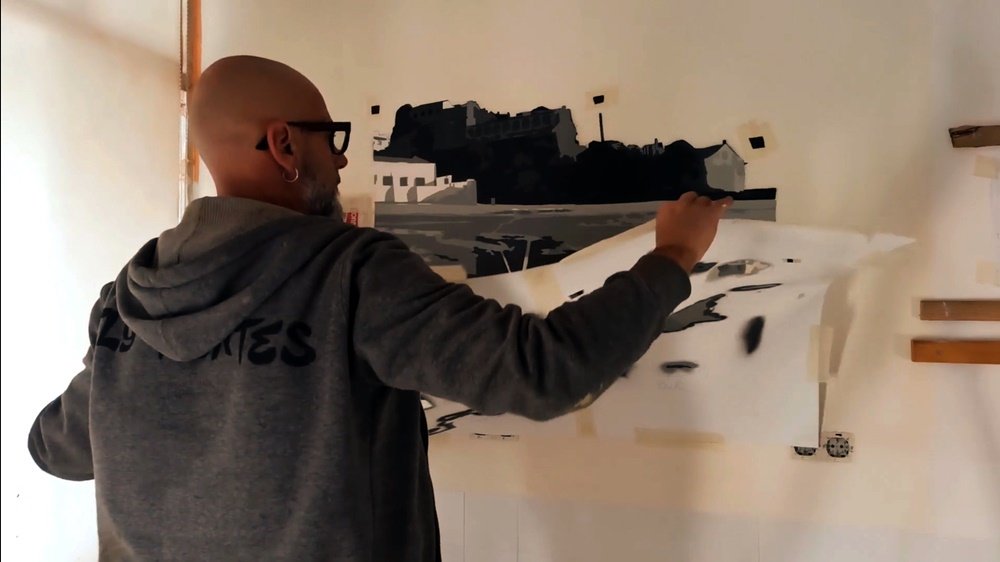O artista Fly Pontes a remover uma das camadas de stencil do Forte de Milfontes no mural. Este passo revela a silhueta e os detalhes arquitectónicos do Forte, construindo a sua forma através da técnica de camadas.