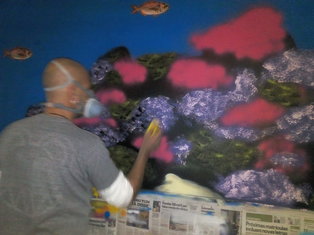 O artista Fly Pontes a pintar o coral no mural "O Ecossistema Marinho Local", utilizando uma esponja para dar textura. Algumas rochas estão quase prontas e outras ainda nas cores iniciais, demonstrando a progressão da pintura.