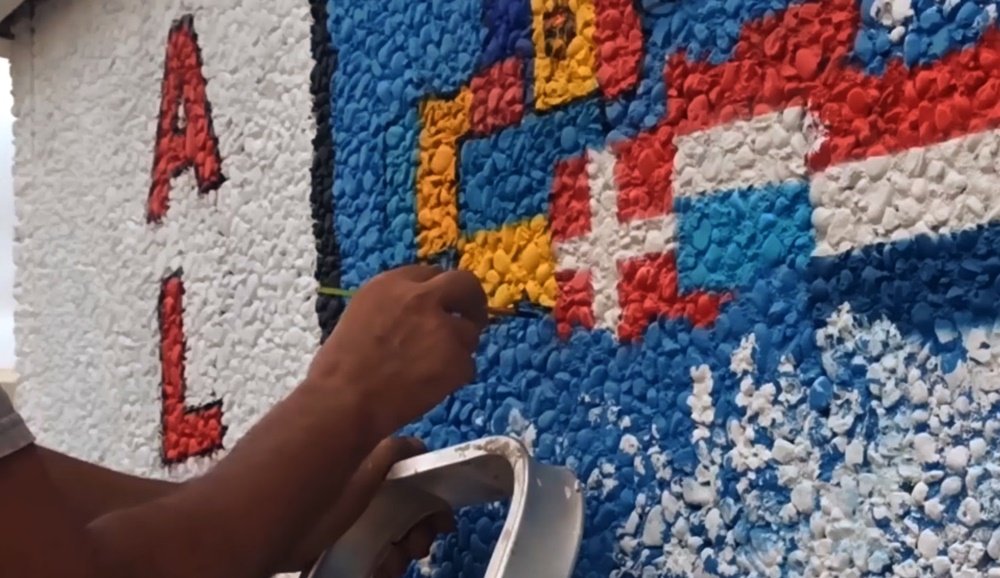 O artista Fly Pontes a pintar uma das muitas bandeiras na lateral do PT de Almancil, dedicada à Diversidade Cultural, utilizando um pincel fino para os detalhes e cores de cada nação.