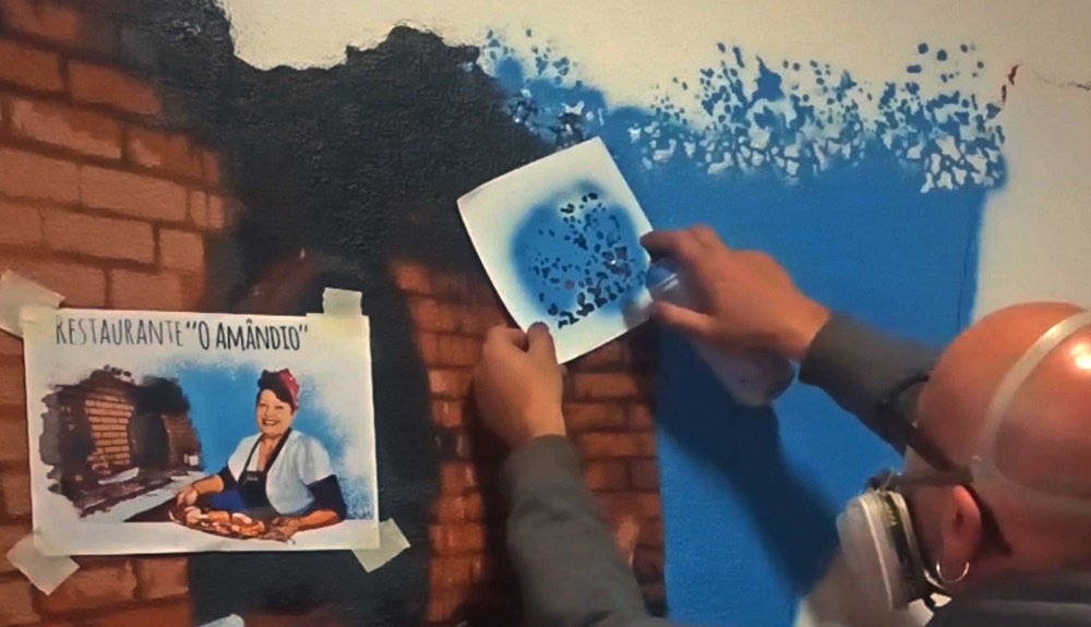 O artista Fly Pontes a pintar retoques de azul sobre os tijolos da grelha já pintada na parede. A folha de referência está visível, guiando a integração e os efeitos de cor para a figura da Julinha.