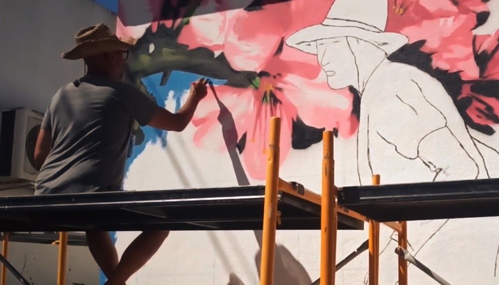 O artista Fly Pontes sentado no andaime, a pintar a última camada de fundo azul no mural. A figura da Marquinhas e os ramos de amendoeira em flor já estão terminados, e o azul realça o forte contraste minimalista.