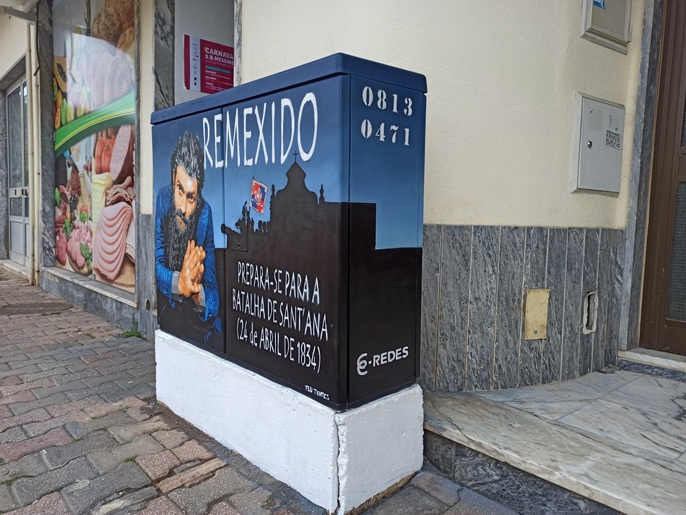 Vista lateral do armário onde se vê o prolongamento da Ermida de Sant'Ana, o palco da histórica Batalha de Sant'Ana. A imagem inclui uma parte da frente, onde está a figura do Remexido, reforçando o contexto histórico do mural em Messines.