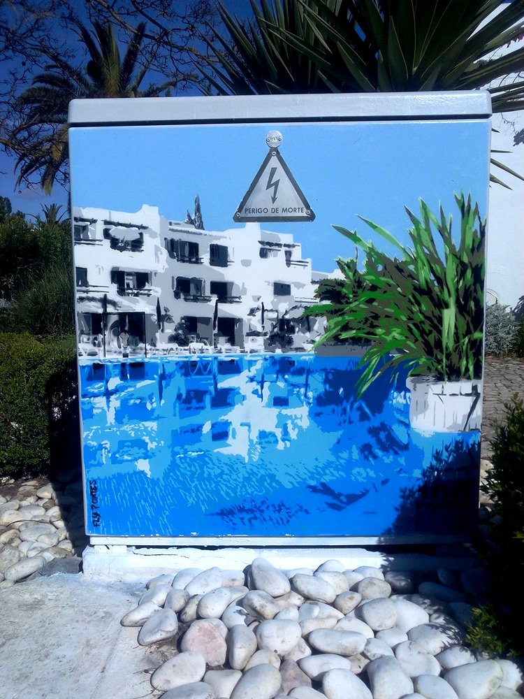 Pintura "Reflexo na Piscina" terminada num armário de electricidade no Hotel da Balaia. O edifício em tons de cinza sóbrio reflete-se na água azul vibrante da piscina. A obra celebra a arquitectura do resort com a técnica Stencil e Spray.