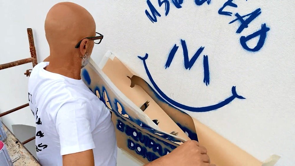 O artista Fly Pontes a retirar a primeira camada do stencil do logotipo da Unidade Sócio-Ocupacional (USO) por cima da entrada do pavilhão. A imagem mostra a aplicação da técnica de stencil para precisão no branding.