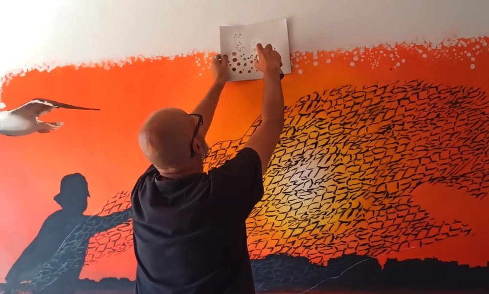 O mural "O Pôr do Sol do Pescador" terminado. O artista Fly Pontes dá os últimos retoques utilizando um stencil com círculos de vários tamanhos para criar uma moldura decorativa à volta da pintura.
