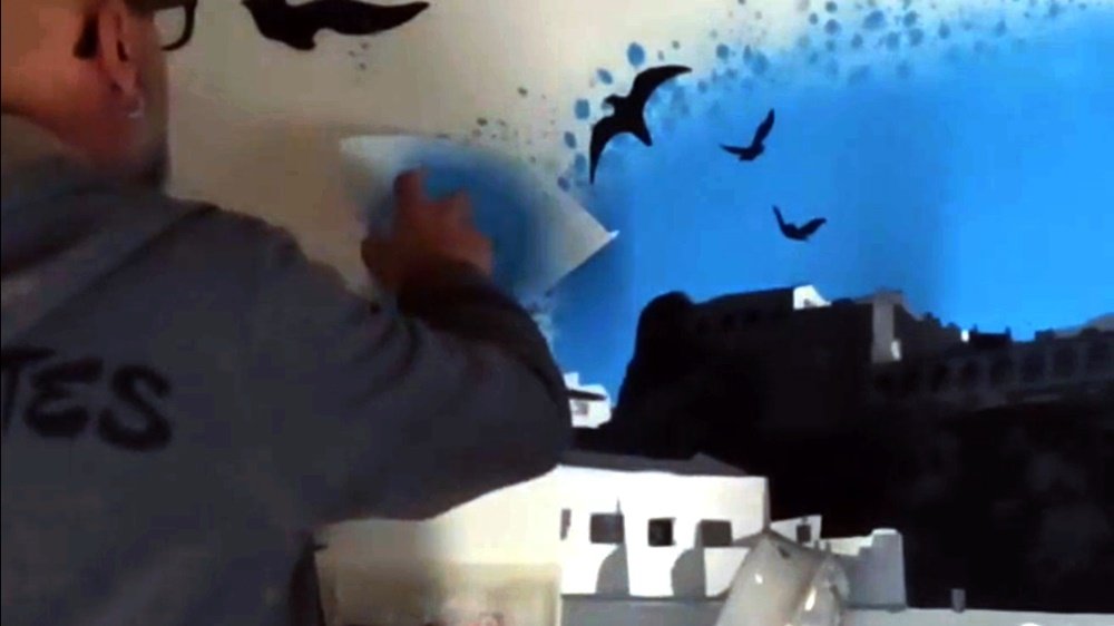 O artista Fly Pontes a pintar os círculos da moldura do mural "Forte de Milfontes" com a técnica de stencil. A vista aproximada mostra parte do Forte e as silhuetas de pássaros a esvoaçar, unindo a arte principal à moldura final.