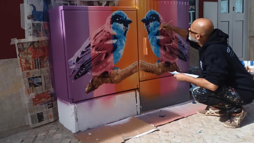 O artista Fly Pontes a dar retoques com pincel fino nos pardais e no galho já terminados. Falta apenas pintar a base do armário de electricidade de branco para concluir o mural "Pintura dos Pardais" em Messines.