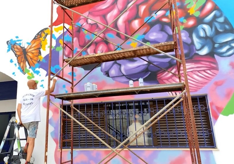 O artista Fly Pontes em cima do escadote a pintar os brancos do mural "A Metamorfose". A pintura (cérebro e borboletas) está quase pronta, e o artista faz retoques para limpar as linhas e definir a imagem da obra para a USO.