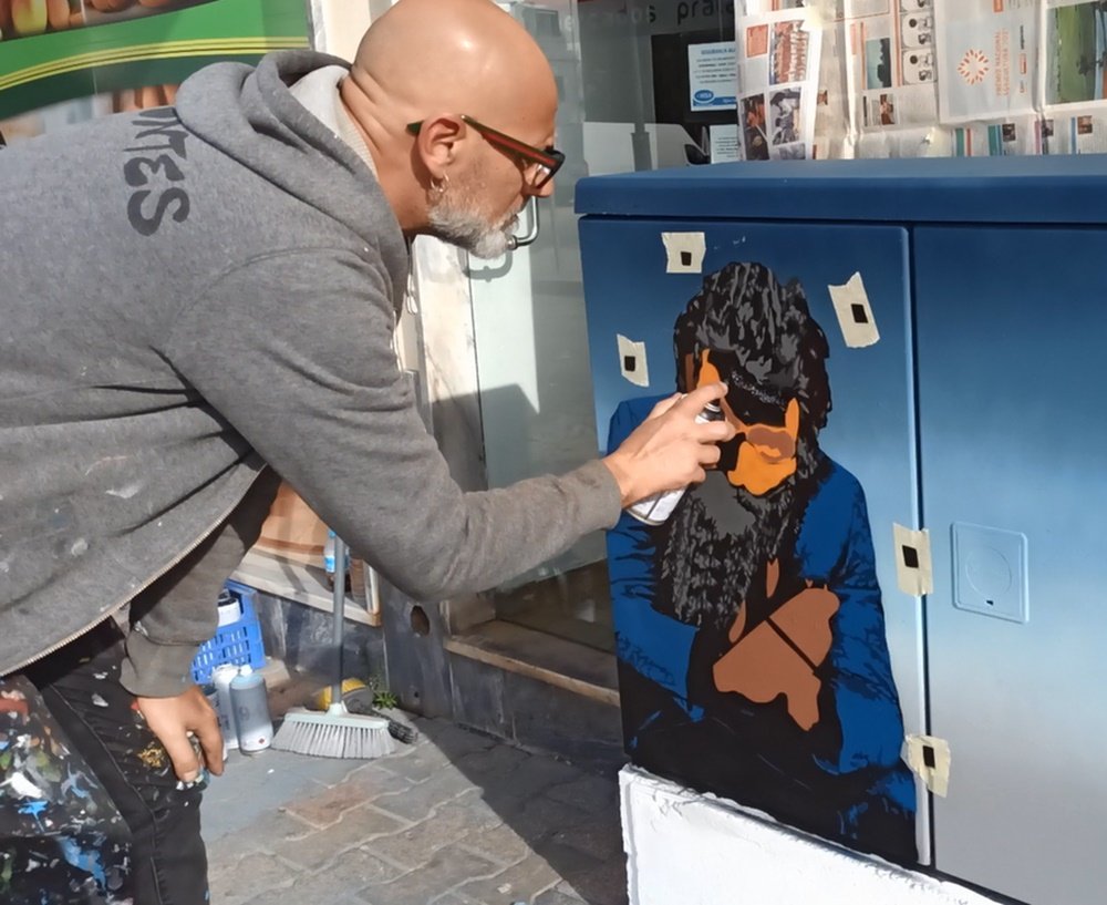 O artista Fly Pontes a aplicar retoques com spray na figura do Remexido. A figura ainda está em fase intermédia. As fitas-cola no armário são visíveis, servindo de marcação para a aplicação do próximo stencil multicamadas.